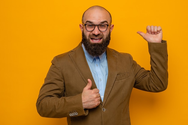 Bearded man in brown suit wearing glasses looking at camera happy and positive showing thumbs up pointing back with thumb standing over orange background