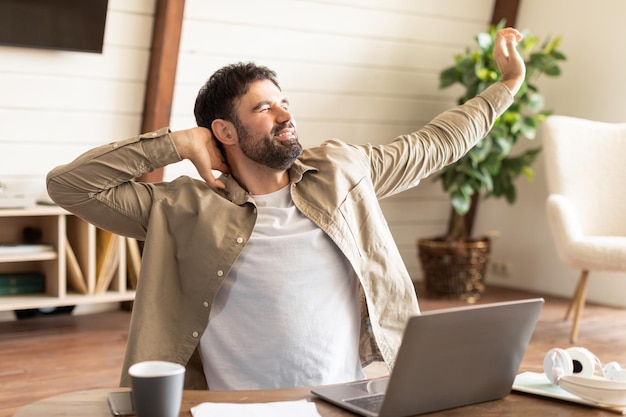 A bearded man in casual attire is stretching with his arms above his head leaning back in his chair