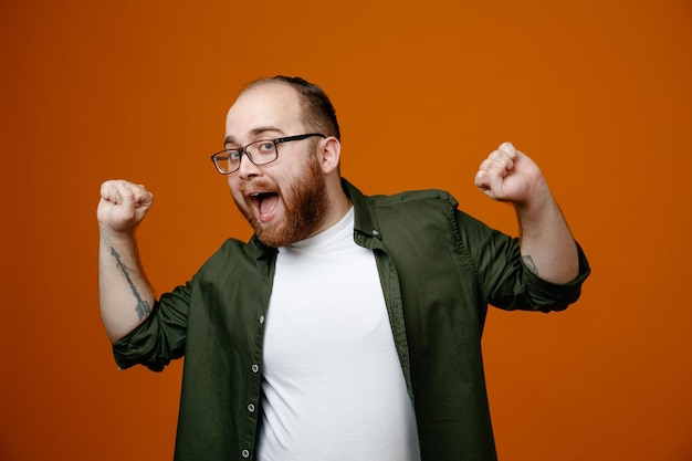 Bearded man in casual clothes clenching fists crazy happy rejoicing his success standing over orange background