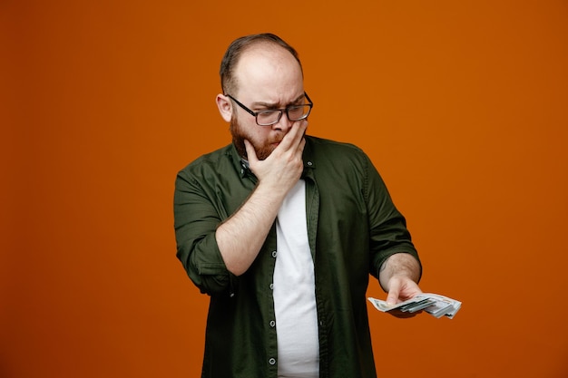 Bearded man in casual clothes wearing glasses holding cash looking puzzled covering mouth with hand thinking standing over orange background