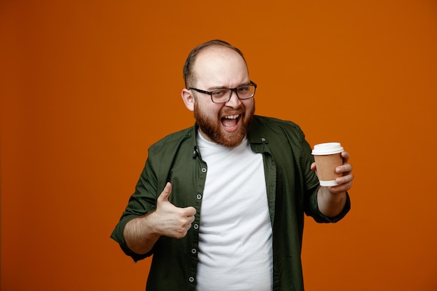 Bearded man in casual clothes wearing glasses holding cup of coffee looking at camera happy and positive smiling broadly showing thumb up standing over orange background