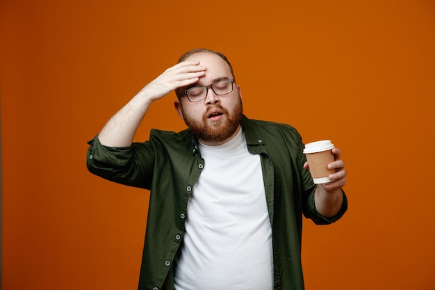 Bearded man in casual clothes wearing glasses holding cup of coffee looking confused holding hand on his forehead for mistake standing over orange background