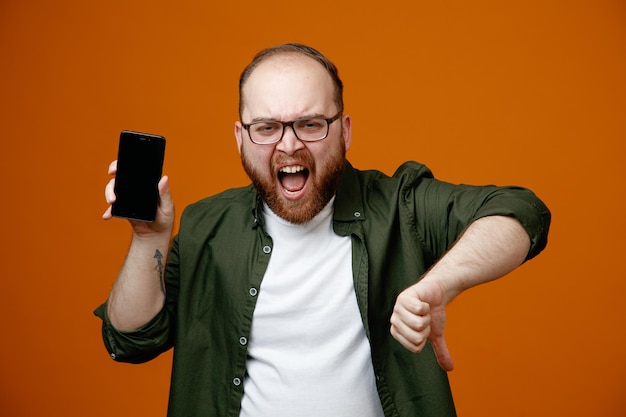 Bearded man in casual clothes wearing glasses holding smartphone looking at camera angry and frustrated showing thumb down standing over orange background