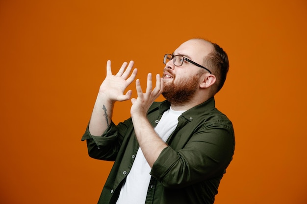 Bearded man in casual clothes wearing glasses looking aside frightening raising arms standing over orange background