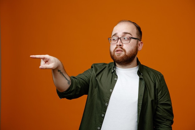 Bearded man in casual clothes wearing glasses looking aside pointing with index finger at something being confused standing over orange background