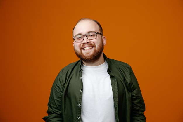 Bearded man in casual clothes wearing glasses looking at camera happy and positive smiling broadly standing over orange background