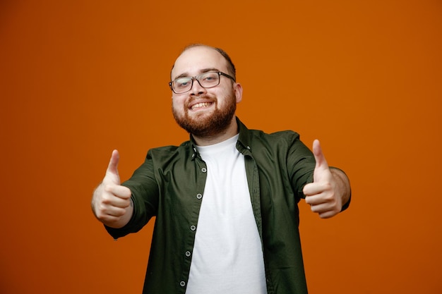 Bearded man in casual clothes wearing glasses looking at camera smiling broadly happy and positive showing thumbs up standing over orange background