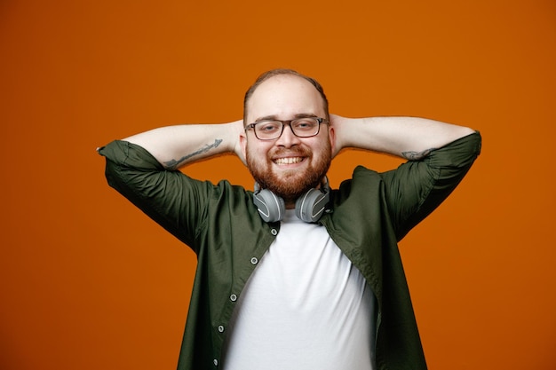 Bearded man in casual clothes wearing glasses with headphones around neck looking at camera happy and relaxed with hands behind his head smiling broadly standing over orange background