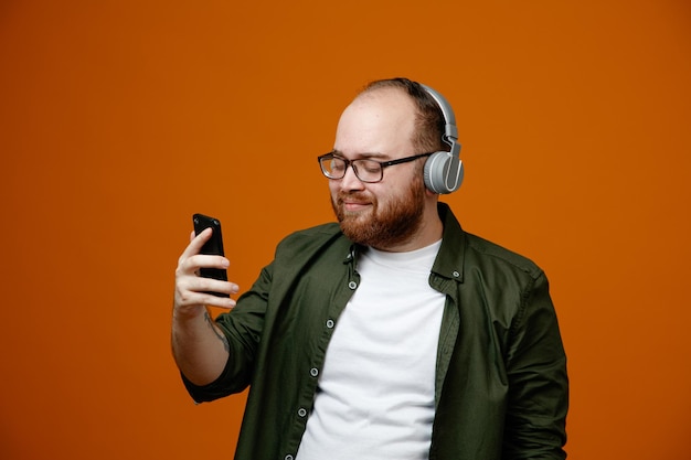 Bearded man in casual clothes wearing glasses with headphones holding smartphone looking at screen with smile on happy face standing over orange background