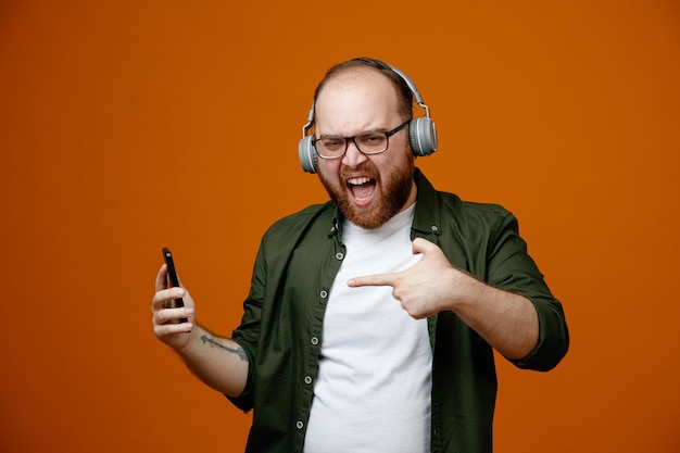 Bearded man in casual clothes wearing glasses with headphones holding smartphone pointing with index finger at it looking excited and happy standing over orange background