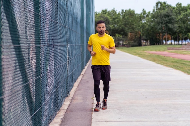 Bearded man in a comfy tracksuit runs along a road passing a high mesh fence on a sports ground