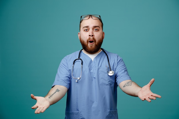 Bearded man doctor in uniform with stethoscope around neck wearing glasses looking at camera happy and surprised spreading arms to the sides standing over blue background