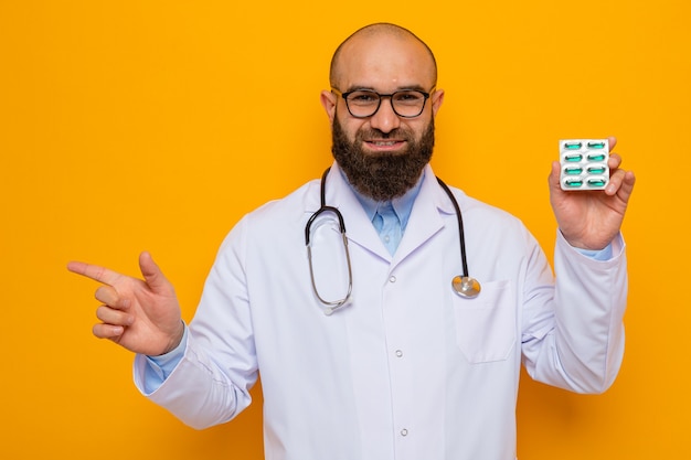 Bearded man doctor in white coat with stethoscope around neck wearing glasses holding blister with pills  happy and positive pointing with index finger to the side