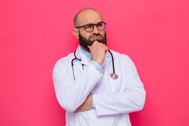 Bearded man doctor in white coat with stethoscope around neck wearing glasses looking aside with hand on his chin thinking with serious face standing over pink background