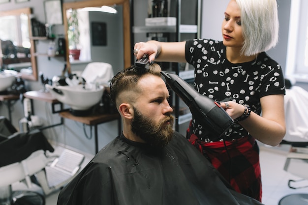 Bearded man at hairdresser with hair dryer while sitting in chair at barbershop.