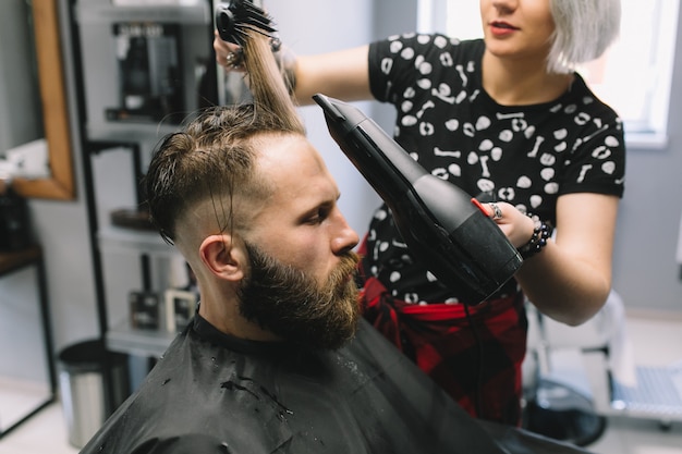 Bearded man at hairdresser with hair dryer while sitting in chair at barbershop.