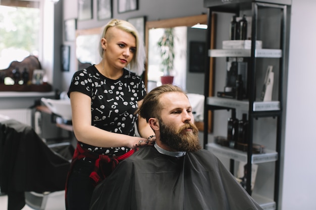 Bearded man at hairdresser with hair dryer while sitting in chair at barbershop.