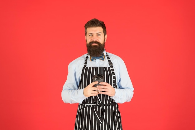 Photo bearded man hold take away coffee. brutal waiter in cafe. mature man red background. skilled servant. barista hipster chef apron. elegant confident bartender. male drink coffee. robusta arabica blend.