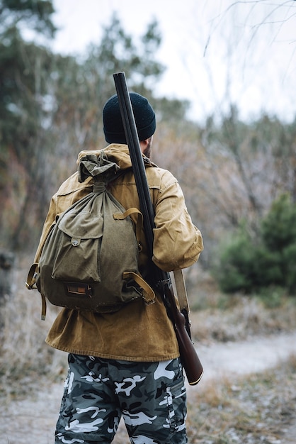 A bearded man hunter in a dark warm hat in a khaki jacket and camouflage pants with a gun on his shoulder