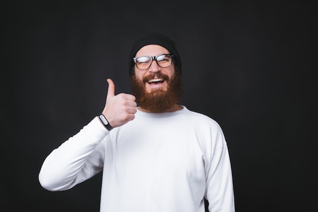 Bearded man  is smiling and showing thumb up on black background.