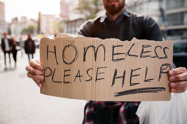 Bearded man is standing on street and holding a cardboard. It says homeless please help. Guy is looking for some mercy from other people and help as well.