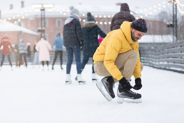 Bearded man in jacket tying shoelaces on ice rink in snowy winter day