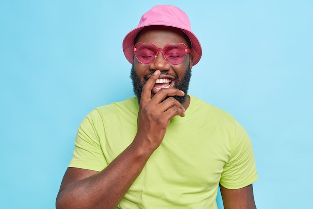 Bearded man laughs happily shows white perfect teeth keeps eyes closed wears pink panama heart shaped sunglasses casual green t shirt expresses positive emotions isolated on blue studio wall