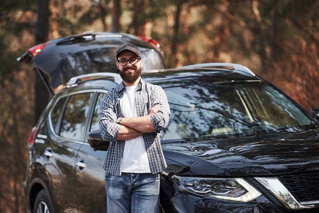 Bearded man near his brand new black car in the forest. Vacations concept