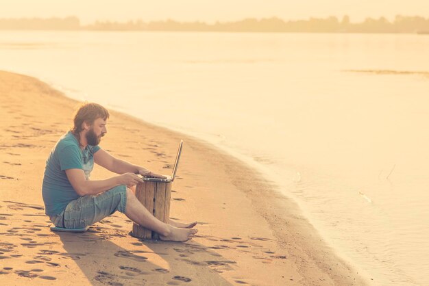 Bearded man in shorts sitting on a sandy beach near water and working at a laptop sunset or sunrise Toned