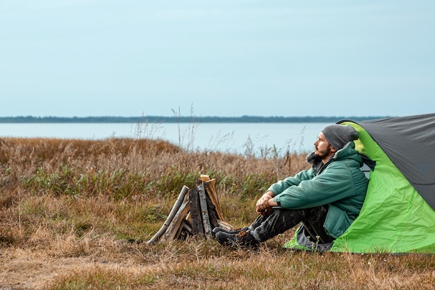 A bearded man sits camping in a tent against the backdrop of nature and the lake.  travel, tourism, camping.