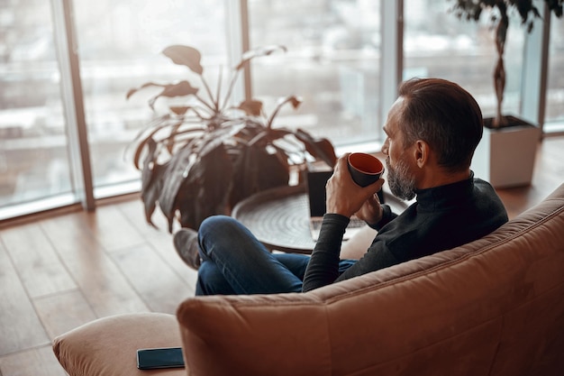 Bearded man sitting in armchair and holding cup of hot drink