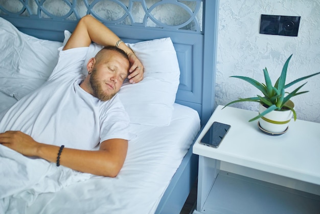 Bearded man sleeping alone on a big and cozy bed white linens with smartphone