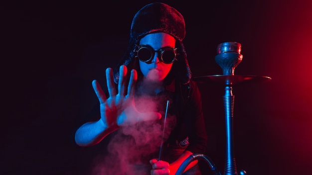 Bearded man smokes a shisha in a hookah bar and blows a cloud of smoke on a dark background with neon lighting