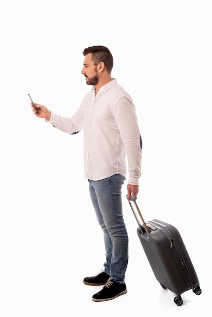 Bearded man standing with a travel suitcase looking at his cell phone on white background