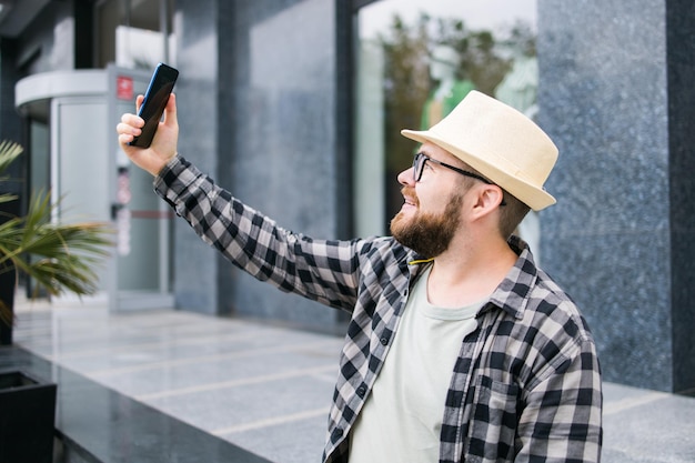 Bearded man takes selfie via smartphone strolls in downtown poses against modern city building enjoys leisure time People urban lifestyle and technology concept