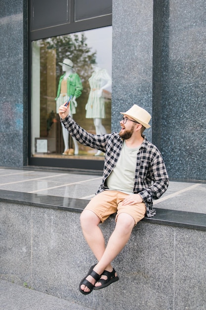 Bearded man takes selfie via smartphone strolls in downtown poses against modern city building enjoys leisure time People urban lifestyle and technology concept