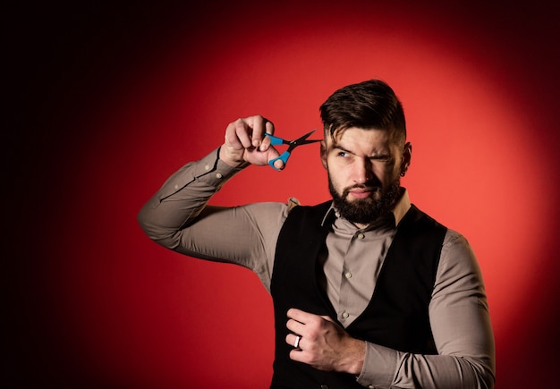 Bearded man in vest on red background. Men cut their hair with hairdressing scissors