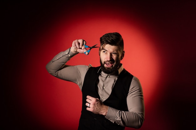 Bearded man in vest on red background. Men cut their hair with hairdressing scissors