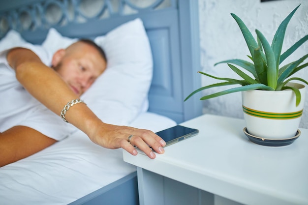 Bearded man waking up on a big and cozy bed with mobile alarm clock woken by