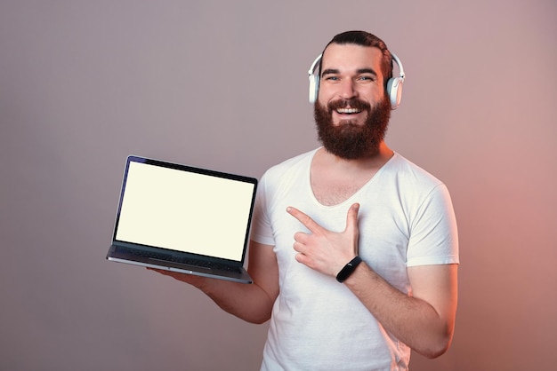 Bearded man wearing earphones is pointing at the blank screen of laptop