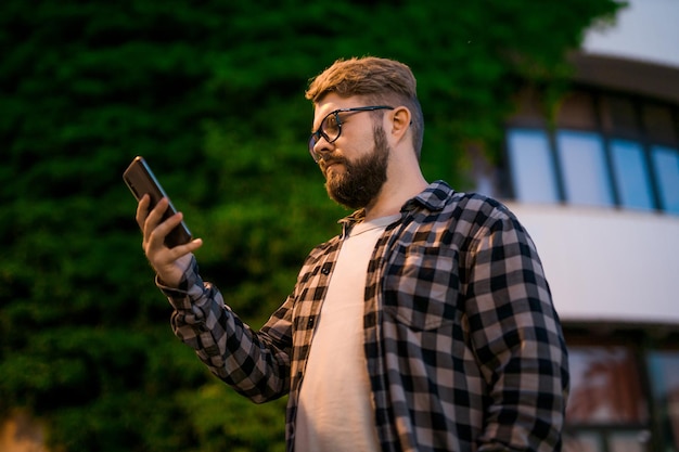 Bearded man wearing eyeglasses is holding scrolling texting in his cellphone at night street guy cal