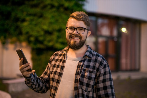 Bearded man wearing eyeglasses is holding scrolling texting in his cellphone at night street guy cal