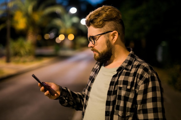 Bearded man wearing eyeglasses is holding scrolling texting in his cellphone at night street guy cal