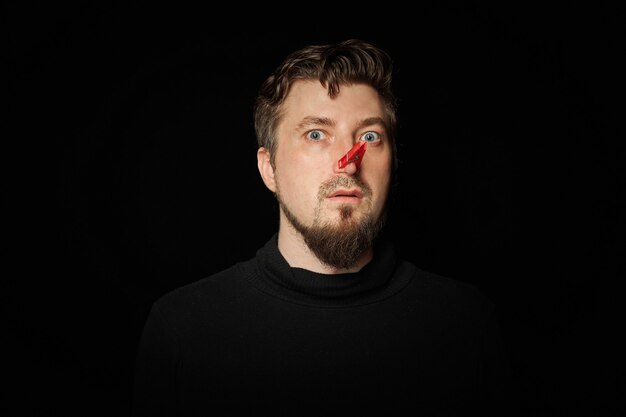 Bearded man with clothespin on nose. Bad stink, funny prankster, joke concept. Laugh at yourself. Red clothespin - symbol of self-mockery and joking character. Black background.