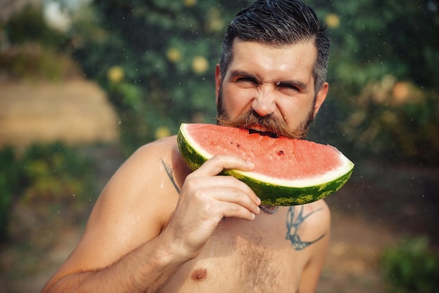 A bearded man with a large red mustache and wet hair holds a slice of ripe red watermelon with his hands, a close-up portrait against the background of a green garden