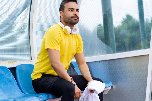 Bearded man with towel and headphones sitting on plastic chair in arbor at sports stadium