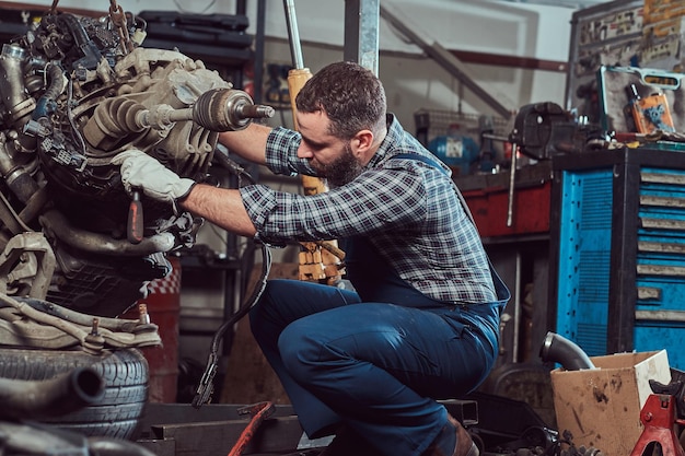 Bearded mechanic specialist repairs the car engine which is raised on the hydraulic lift in the garage. Service station.