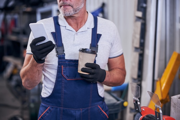 Bearded mechanic using cellphone and holding cup of coffee