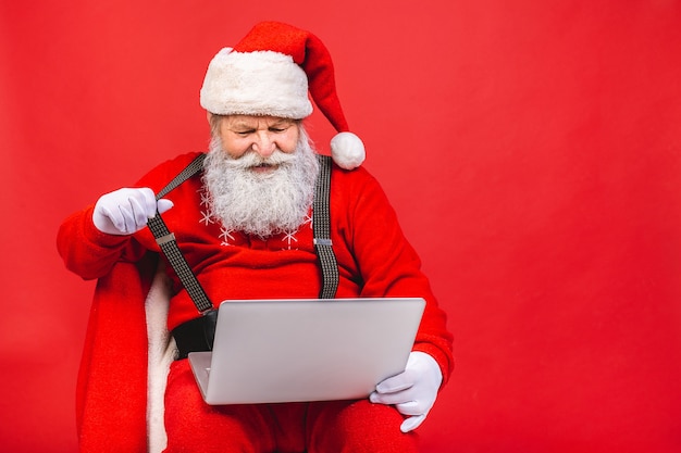 bearded old man in santa costime holding a laptop isolated on red background