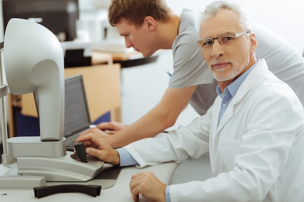 Bearded ophthalmologist. Grey-haired bearded ophthalmologist wearing uniform sitting near his assistant